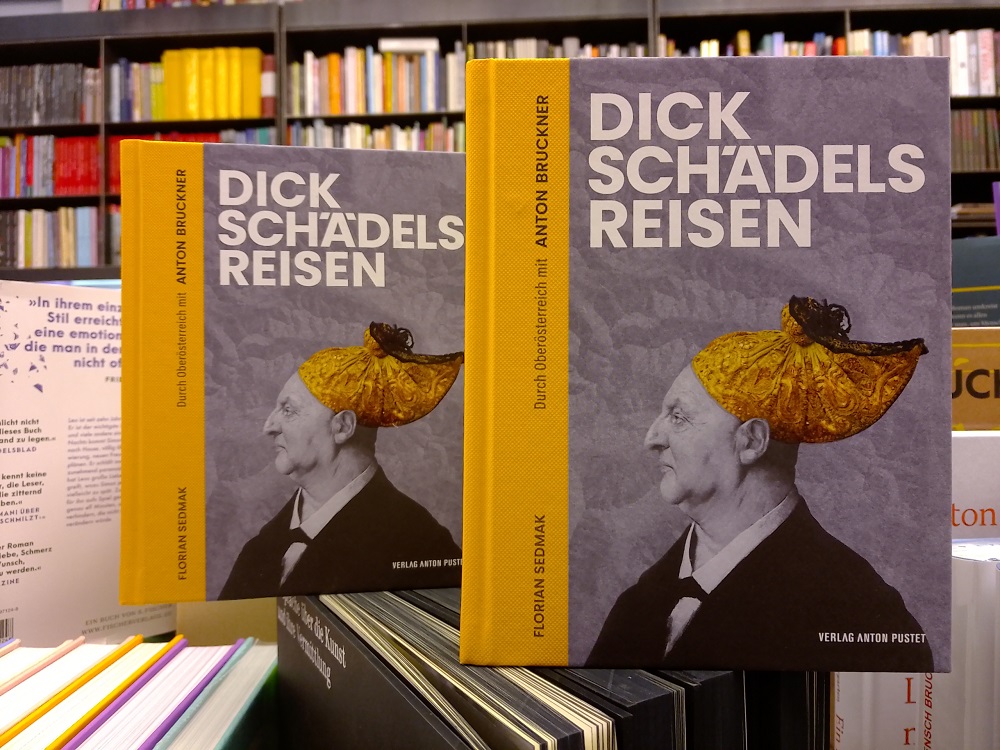 zwei Exempalre des Buches "Dickschädels Reisen" von Florian Sedmak: das Buch hat einen dottergelben Rücken aus strukturiertem Papier, auf dem Cover ist ein Seitenprofil-Foto von Anton Bruckner in schwarz-weiß, auf seinem Kopf trägt einer eine Goldhaube